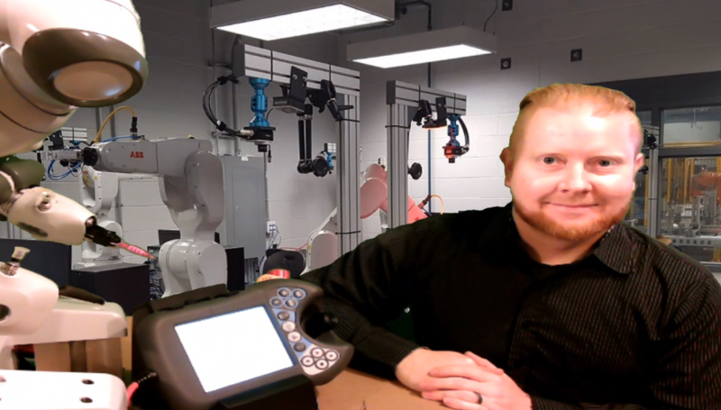 Thomas Forget delivers online lectures from his garage with a photo of one of St. Clair College’s robotics labs behind him. Next to him is the ABB YuMi robot he uses to demonstrate. 